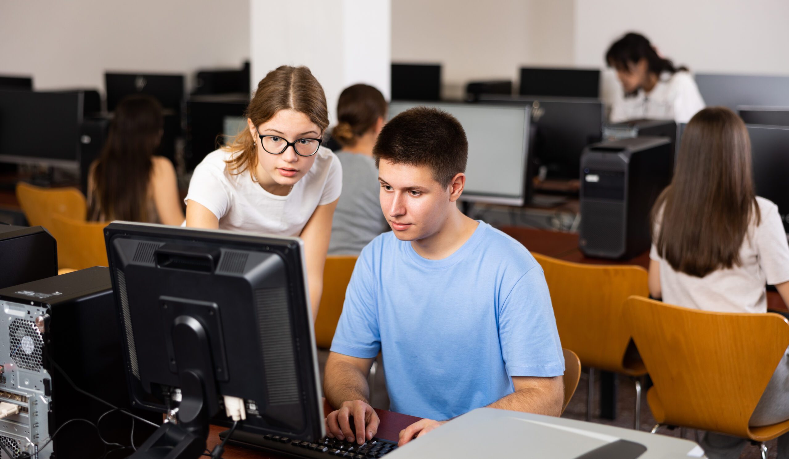 Clever female teenager helping to the male classmate while he is learning computer science in the class